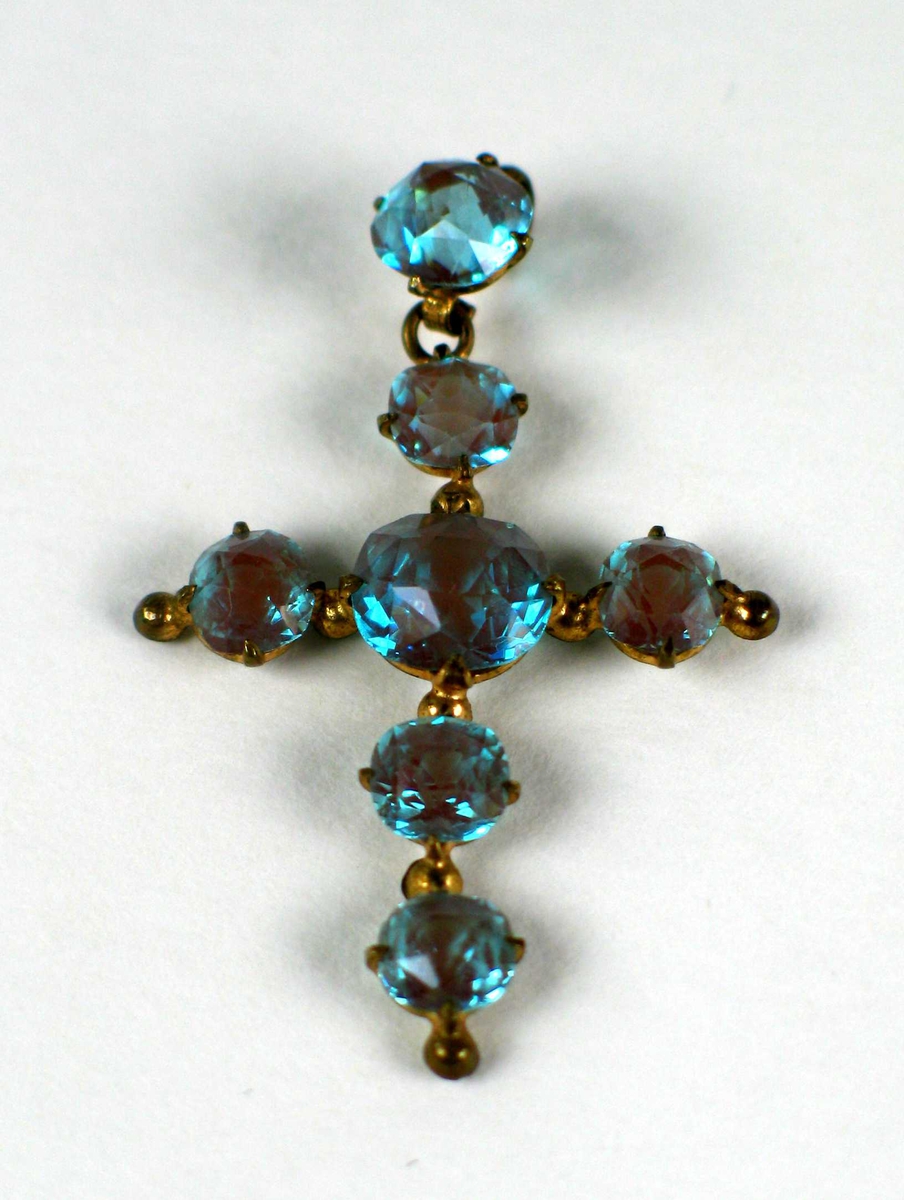 Korsformet anheng antatt i gull med syv slipte turkise steiner. Smykket ligger i et svart etui.