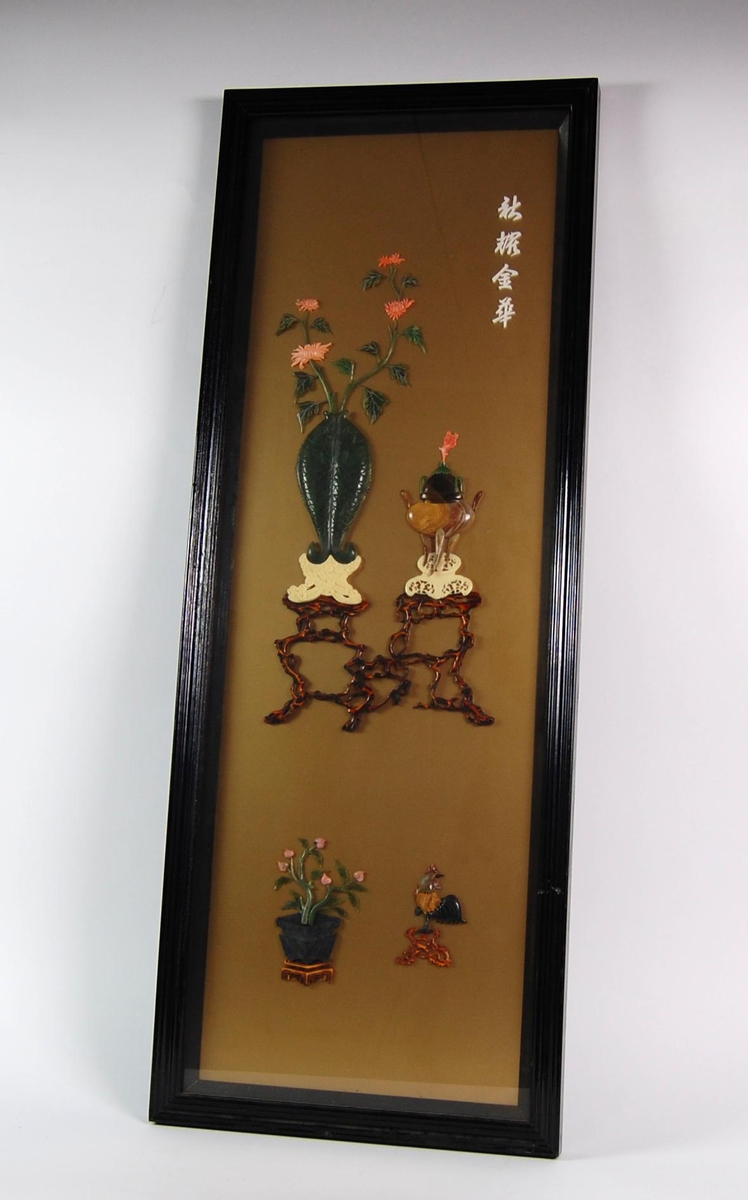 Innrammet bilde med motiv av blomster i vase og en kjele som står på bord. Det er også motiv av en blomsterpotte og en fugl. Motivet er håndlaget av stein og koraller. Rammen og bakplaten er av tre og det er et deksel av glass. Opphengshempen er av metall.