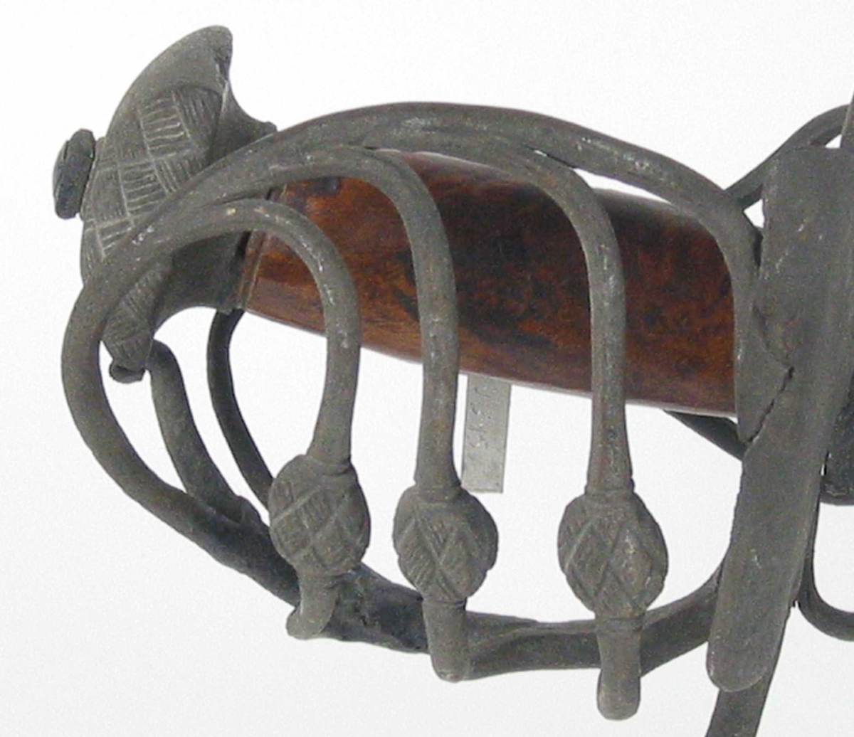 Pyramideformet knapp med kryssfilt dekor og asymmetrisk feste med 3 sidebøyler med fortykninger på midten på utsiden og en på innsiden. Nyreformet parerplate på utsiden og klingebrekker og tommelbøyle på innsiden. S-formede, runde parérstenger med fortykning med filt dekor og dråpeformet avslutning.
Tregrep. Enegget krum, kileformet klinge med en ryggstillt blodrand. Ryggslipt mot spissen. Spiss brukket. Tre hull på spiss. Ett smedstempel.

