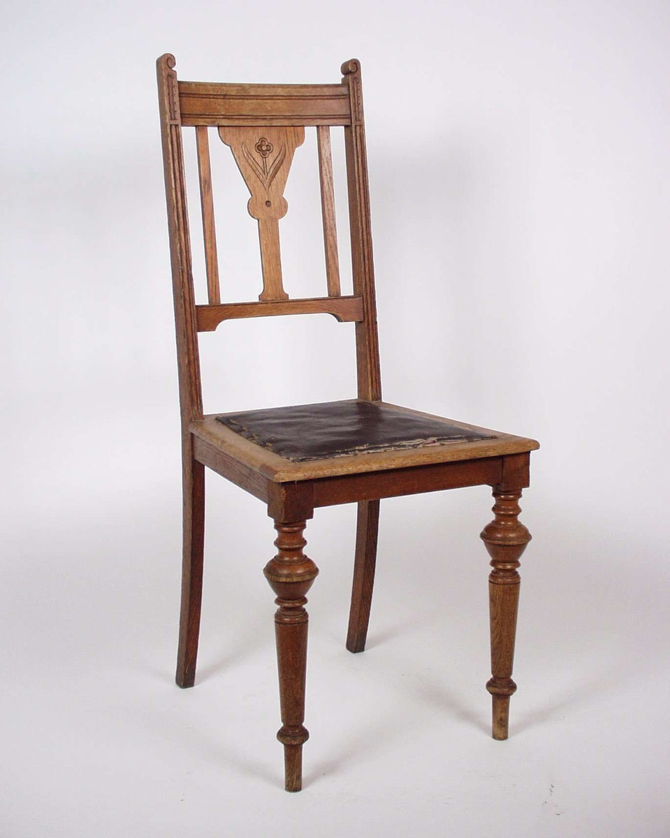 Stolen er av eik med dreide bein og utskjæringer i ryggbrett og toppstykke. Setet er trukket med skinnimitasjon.