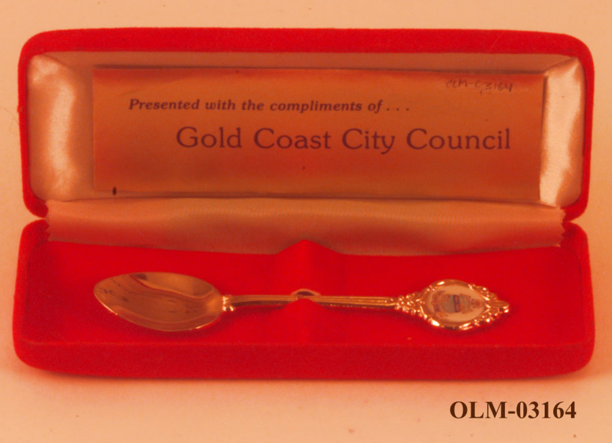 Gullfarget teskje med motiv av et våpenskjold som antageligvis er byvåpenet til Gold Coast. Det følger med en rød eske.