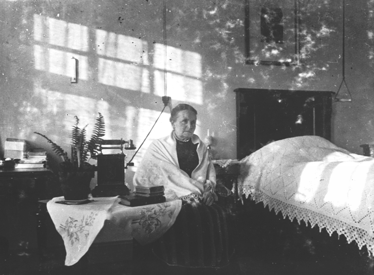 Interiør, soverom. Lisbeth Grüner (1847-1918) ved senga. Telefon på bord. Granerud gård, Brumunddal.