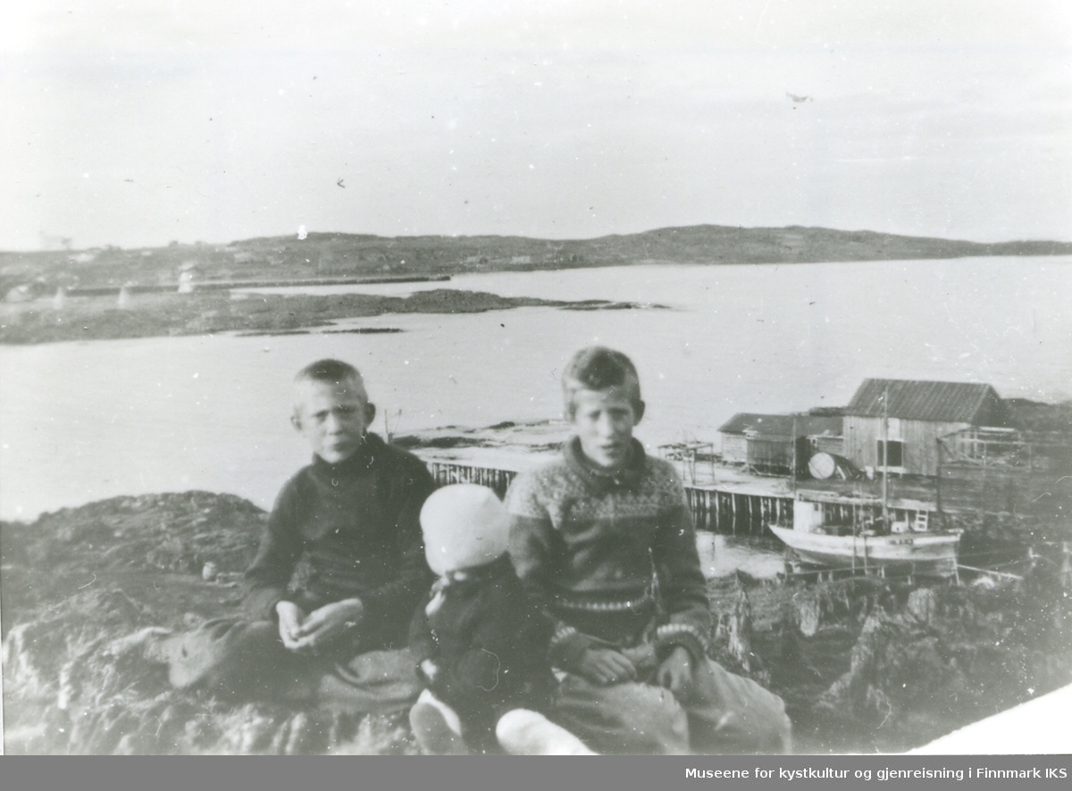 fra v.: Ottar Olsen og søsknene Bjørnar og Odd Olsen sitter på en bergknaus, fiskebruket på sydsiden av Kåven i Gamvik i bakgrunnen.