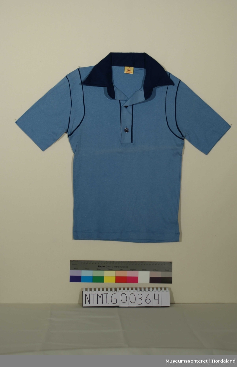 Lyseblå t-skjorte med mørkeblå krave, knappeåpning i hals med to knapper, mørkeblå pyntekanter ved halsåpning og ved ermefeste.