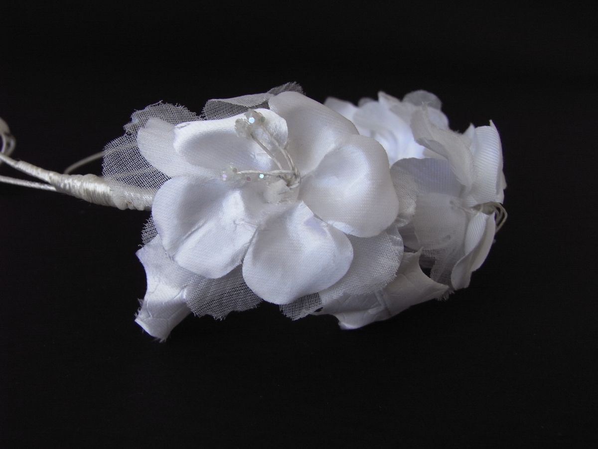 Tre hvite blomster- og bladrosetter på trådomspunnet wire. Blomstene er stiliserte silkeblomster, hver utstyrt med ett dekkblad, og består av tre lag kronblader omkring fem støvdragere prydet med pålimt glitter. Hvert lag består av fem kronblader; de innerste små og dråpeformet av sateng, de mellomste større og hjerteformet av nettstoff og de ytterste hjerteformet av kreppet sateng. Wirekonstruksjonen av millimetertynn metalltråd, er omspunnet med blank silketråd. Mens endestykkene er formet som åpne løkker av enkel tråd, er metalltråden er dobbel der blomstene er festet.