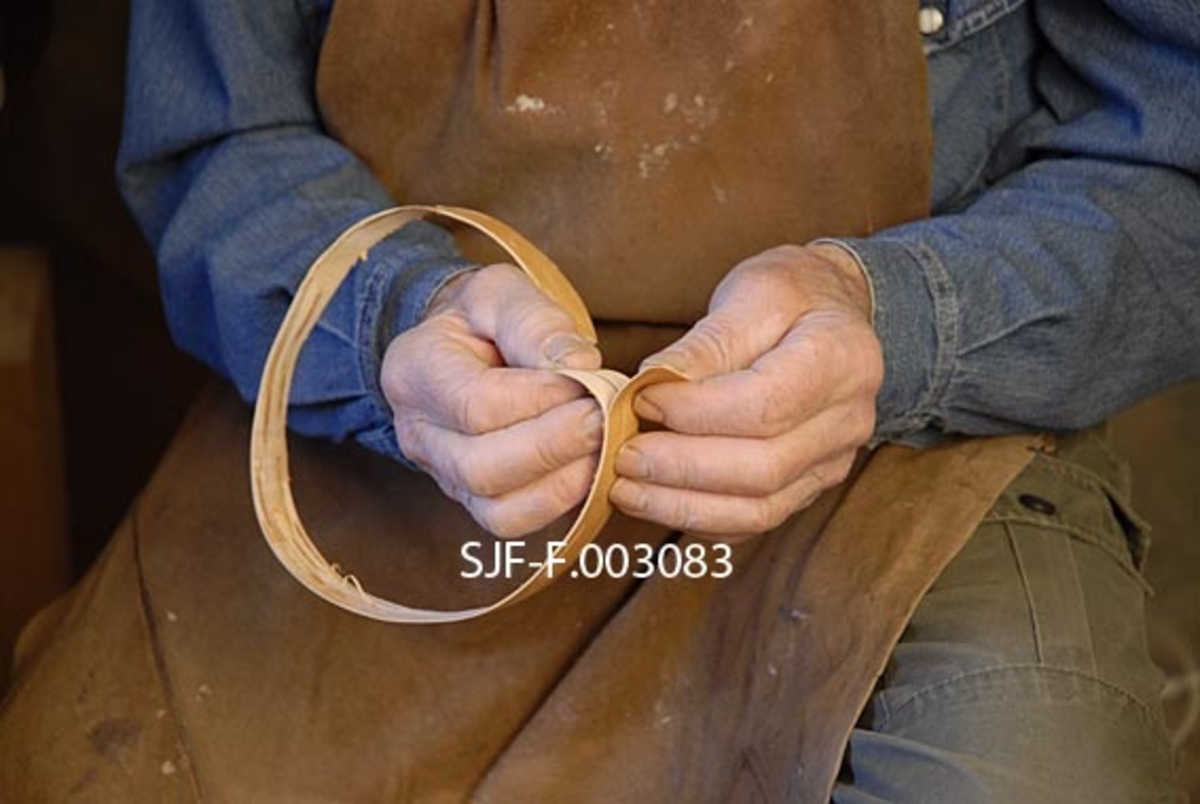 Trygve Løvseth fra Åsnes i Hedmark deler ei tjukk neverremse for å få den tynnere. Dette er et nærbilde av hendene hans og arbeidsstykket. 
Fotografiet inngår i en serie med nummer SJF-F. 003071-SJF-F. 003328.