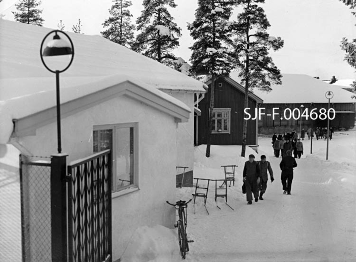 Fra porten til Drammenselvens Papirfabrikker på Modum i Buskerud.  Fotografiet er tatt ved arbeidstidas slutt en marsdag i 1957.  Vi ser en del av fabrikkens ansatte på vanding mot porten.  Til venstre i forgrunnen ligger den kvitmalte murbygningen fra 1954, som inneholdt bad og vasker for de ansatte og deres familier og kontor for portvakta.  Vi ser også at det var ei spesiell «sluse» foran portvaktrommet, der det hang et stemplingsur som mange av de ansatte måtte bortom på veg inn eller (som her) ut av fabrikken.  Ved den nevnte bygningen, like innenfor porten sto det en sykkel og fem sparkstøttinger.  Bak og til høyre for den kvite murbygningen ser vi gavlen på en panelt trebygning som lenge ble brukt som spisebrakke for fabrikkens ansatte, seinere som elektrokontor.  Bak denne igjen sto den store papirlagerbygningen med skyveporter mot jernbanesporet, som gikk langs langveggen mot innkjøringsvegen til fabrikken og elva.  Da dette fotografiet ble tatt sto ei av jernbanens godsvogner oppstilt foran lagerbygningen, antakelig klar for innlasting av papirruller. 