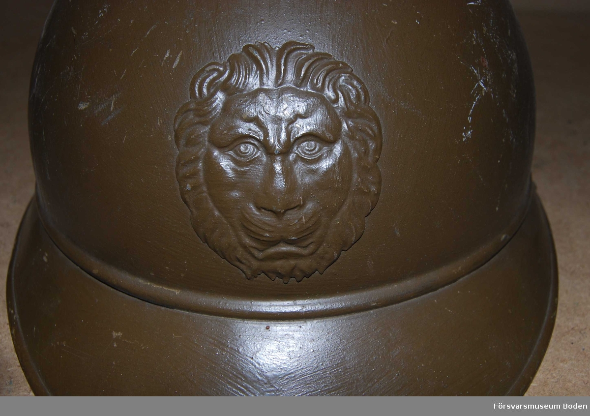 Gulbrun stålhjäm av fransk typ med hakband men utan inredning. Med ett lejonhuvud framtill.