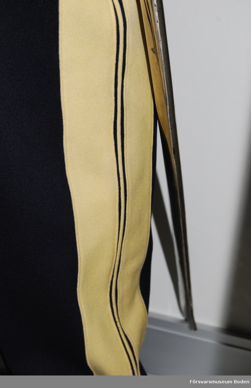 Mörkblått kläde med dubbla breda band av gult kläde (nu blekt) längs yttersömmarna. I centrum en gul passpoal. Dubbla knappar för montering av hällor på byxbenens insidor. Inga hällor är ditsatta.