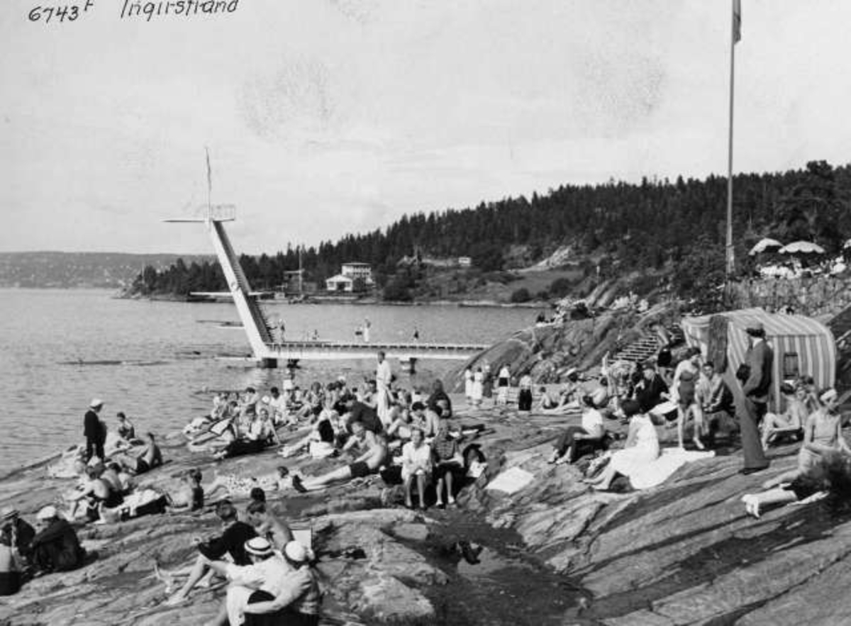 Strandliv med badegjester som soler seg på svaberget ved sjøen på Ingierstrand bad utenfor Oslo. Stupetårn i bakgrunnen.