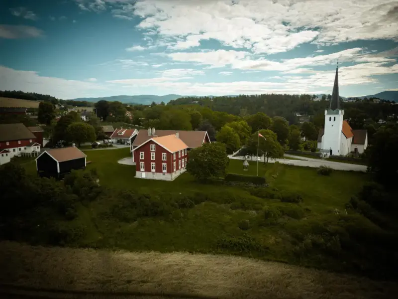 Dronebilde som viser Bildet viser Ringerikes Museums hovedbygg, et brunt stabbur, og Norderhov kirke - en hvitkalket middelalderkirke i stein med høyt mørkegrønt spir. 