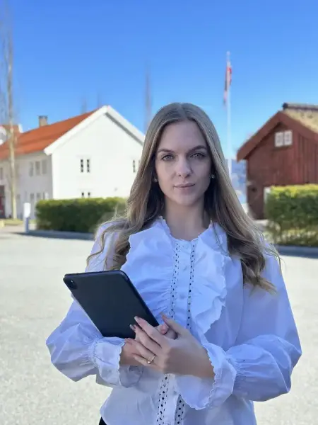 Portrett av en ung kvinne med langt lyst hår og hvit rysjebluse. Holder en ipad. Hus i bakgrunnen.